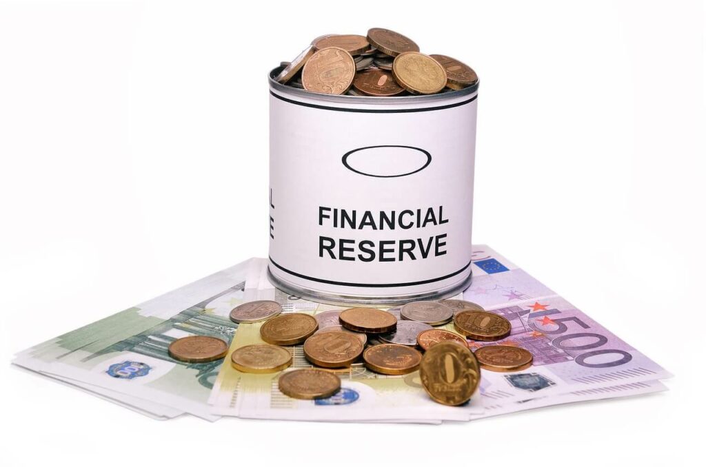 Barattolo di metallo con scritto "FINANCIAL RESERVE" pieno di monete, appoggiato su banconote in euro.