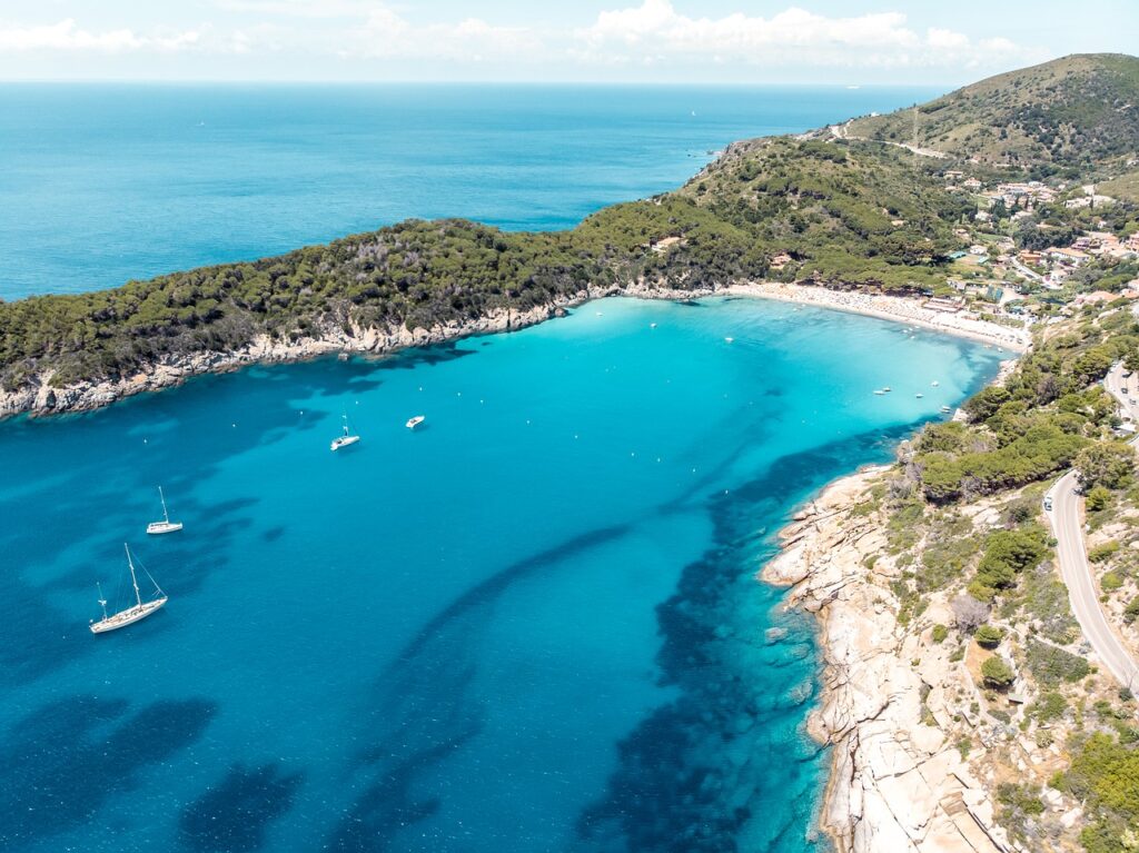 Baia con acqua cristallina circondata da colline verdi e scogliere. Barche a vela ancorate e spiaggia attrezzata sullo sfondo.