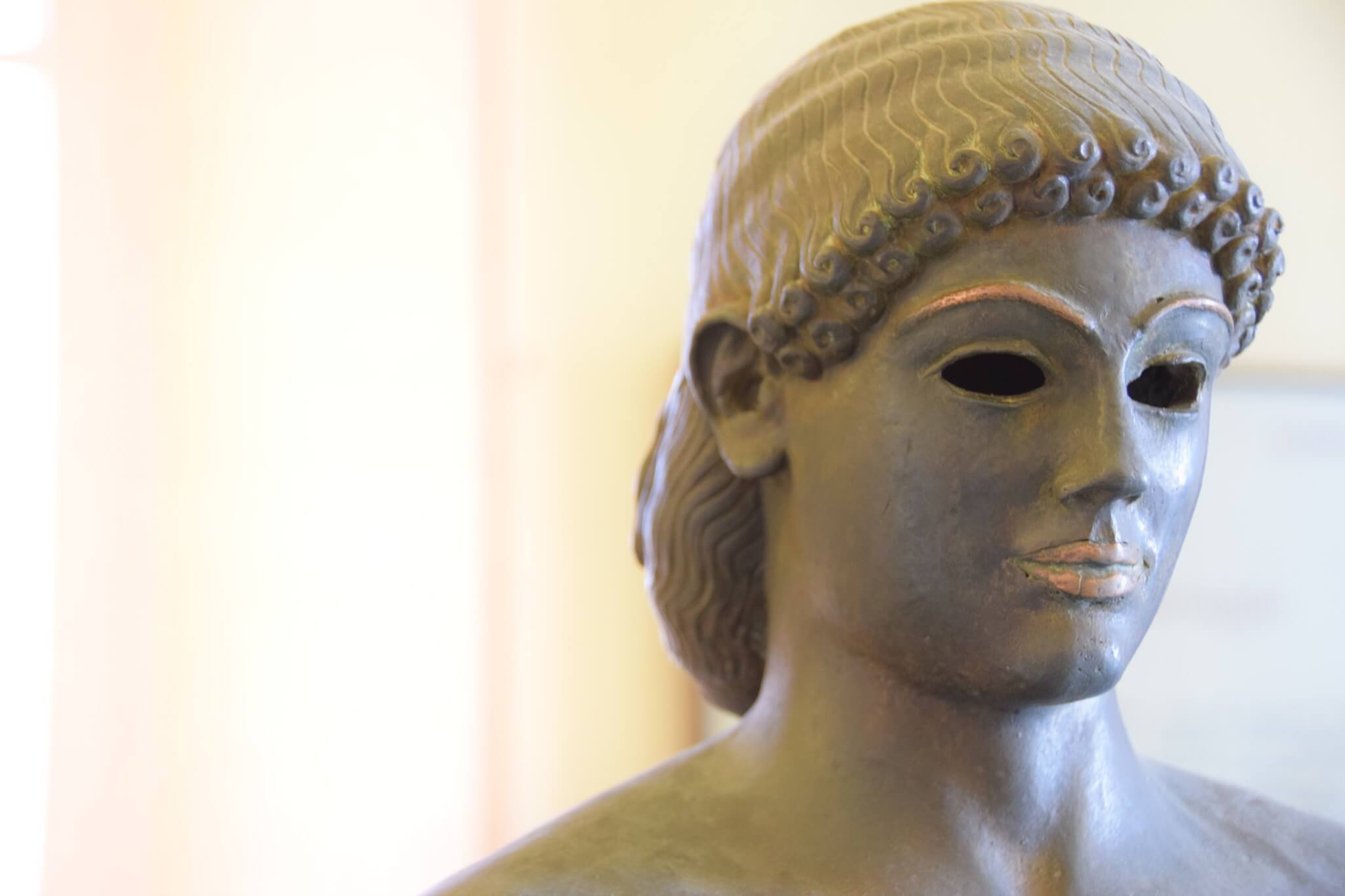 Primo piano di una scultura antica di una testa di donna, realizzata in bronzo scuro con dettagli elaborati nei capelli e nel viso.