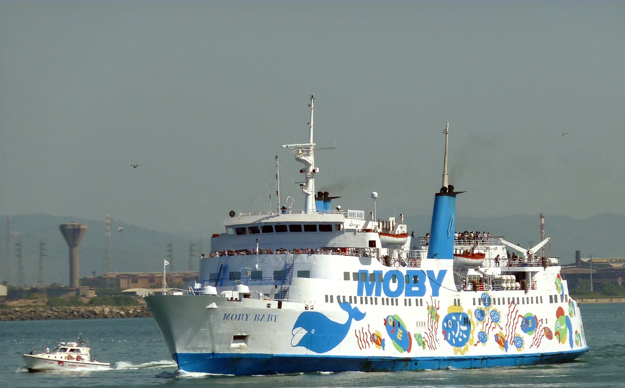 Nave Moby colorata in partenza dal porto di Piombino, con passeggeri a bordo e piccole imbarcazioni nelle vicinanze.