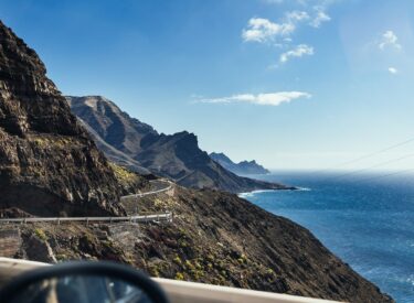 Panoramica di una strada costiera che serpeggia tra montagne e mare, con cielo azzurro e nuvole.