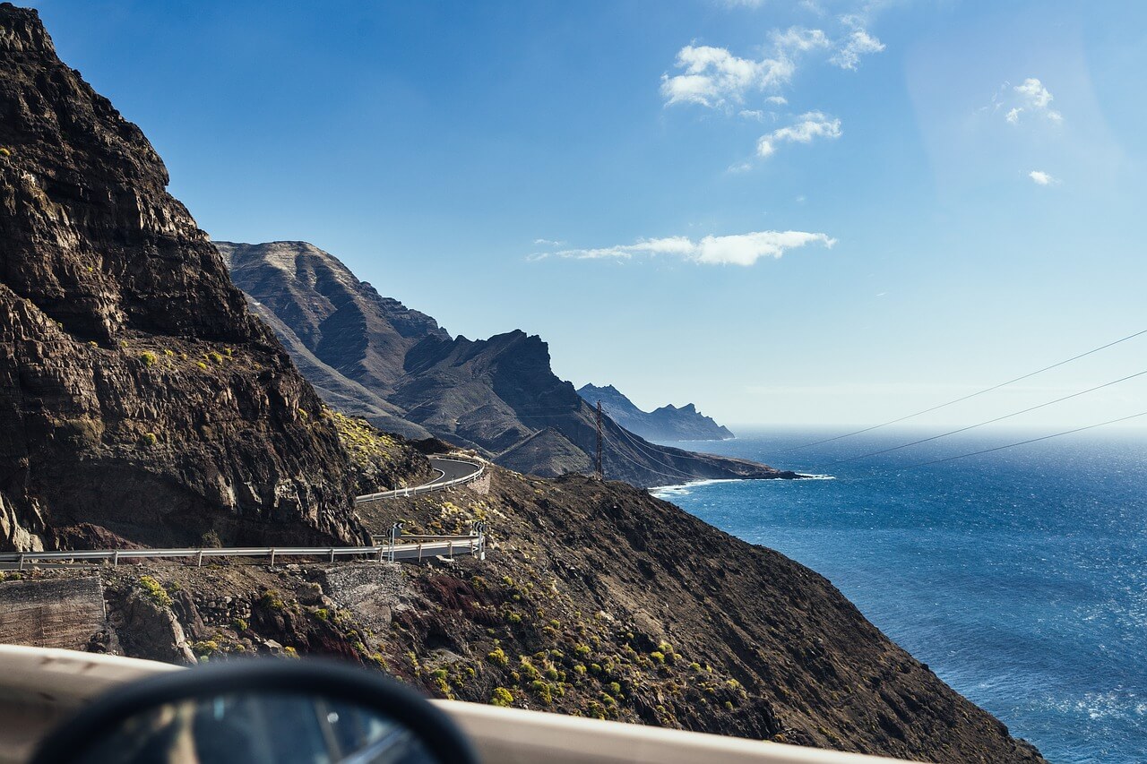 Panoramica di una strada costiera che serpeggia tra montagne e mare, con cielo azzurro e nuvole.