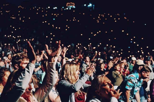 Grande folla di persone applaude ad un evento, con luci soffuse sullo sfondo.