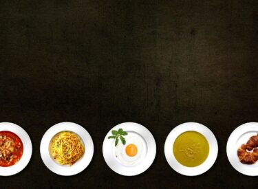 Varie pietanze in piatti bianchi: zuppa, spaghetti, uovo, salsa e cornetti, presentati su sfondo scuro.