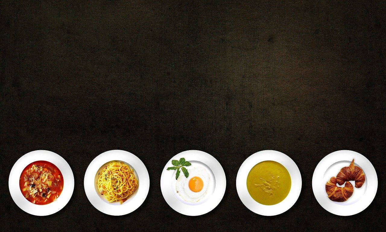 Varie pietanze in piatti bianchi: zuppa, spaghetti, uovo, salsa e cornetti, presentati su sfondo scuro.