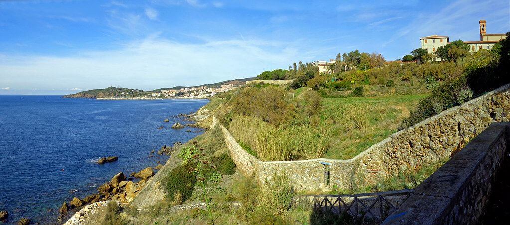 Panorama costiero piombinese con scogliere rocciose, vegetazione e un antico muro di pietra.