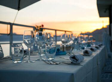 Tavola apparecchiata con bicchieri e posate, con sfondo di tramonto sul mare.