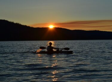 Persona in kayak che pagaia su un lago al tramonto, con il sole che si riflette sull'acqua e colline sullo sfondo.