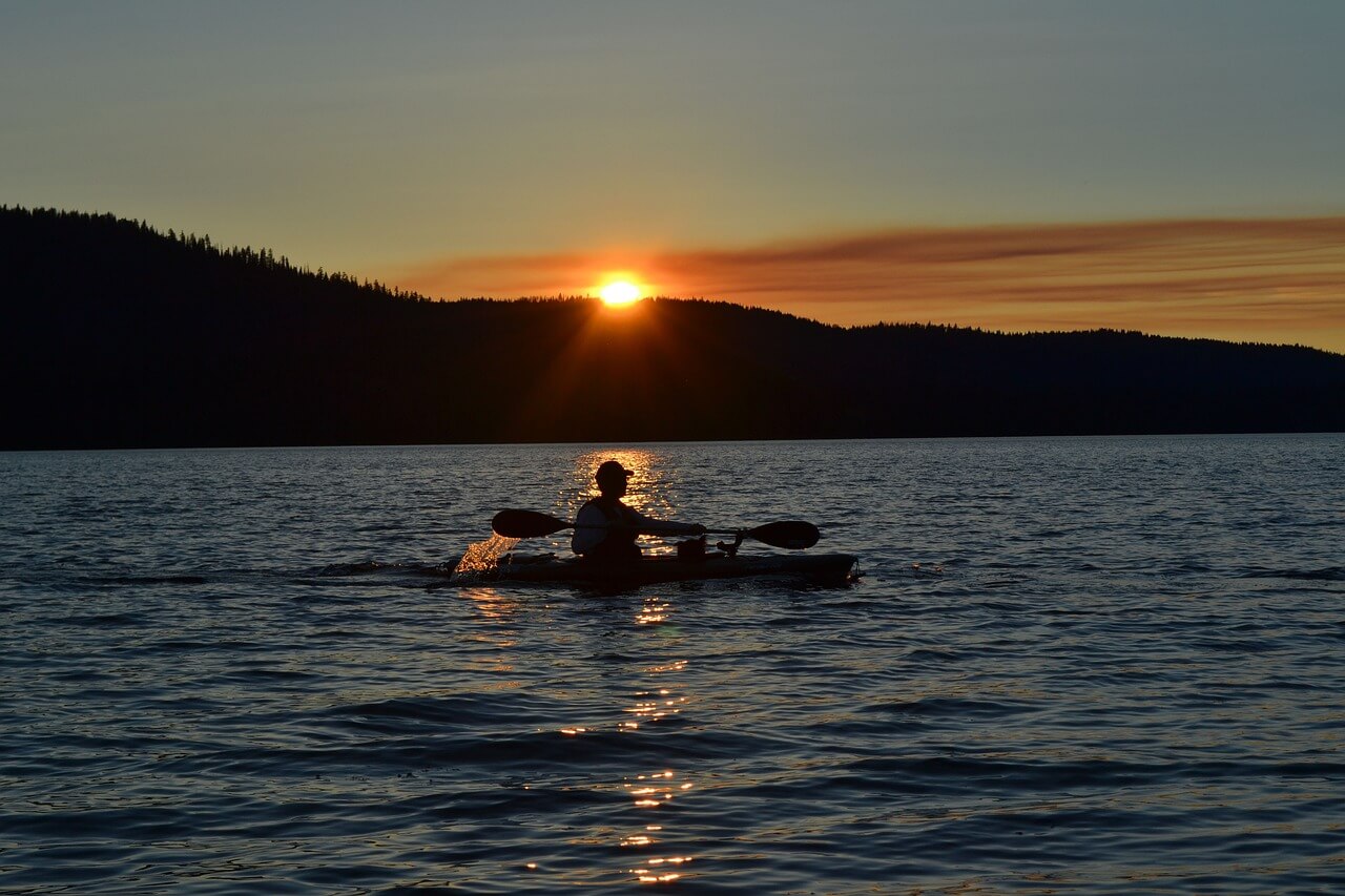 Persona in kayak che pagaia su un lago al tramonto, con il sole che si riflette sull'acqua e colline sullo sfondo.