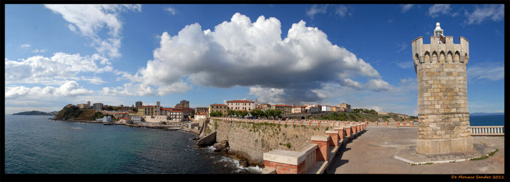 Panorama di Piombino con vista sulla fortezza, il mare e il lungomare. Il cielo è azzurro con grandi nuvole bianche sopra la città.