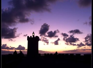 Sagoma di un faro illuminato al tramonto con cielo viola e nuvole sparse, persone in controluce sulla terrazza panoramica vista mare.
