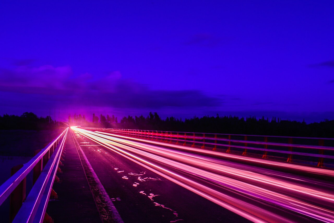 Scia luminosa di auto in movimento su una strada di notte, con un cielo viola scuro sullo sfondo.