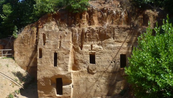Facciata di antiche tombe scavate nella roccia in Toscana, con diverse aperture e vegetazione circostante.