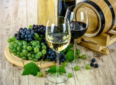 Bicchieri di vino bianco e rosso con grappoli d'uva verdi e viola su un tavolo di legno, vicino a un barile di legno.