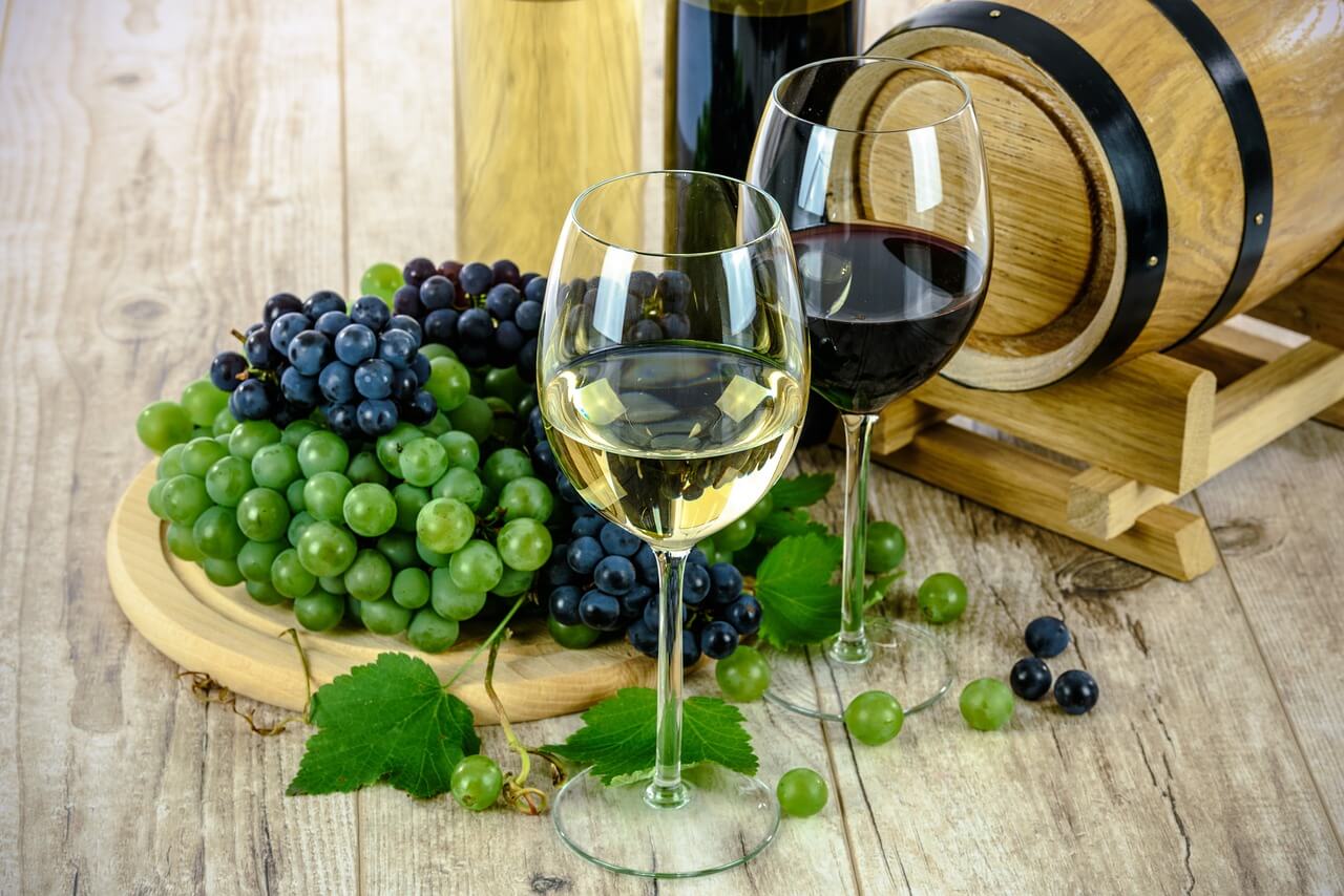 Bicchieri di vino bianco e rosso con grappoli d'uva verdi e viola su un tavolo di legno, vicino a un barile di legno.