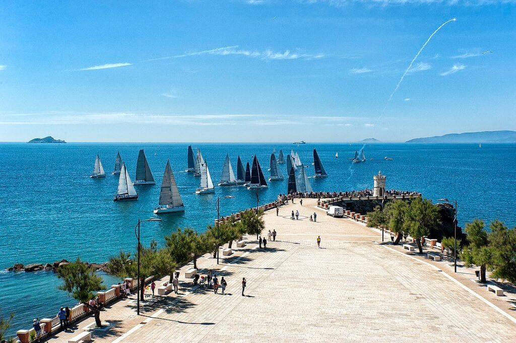 Regata di barche a vela nel mare della Toscana, con velisti in gara e pubblico lungo la passeggiata panoramica vicino al faro.