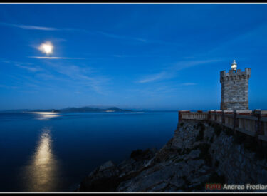Veduta notturna di un faro su una scogliera illuminato dalla luna, con il mare calmo riflettendo la luce e un cielo azzurro scuro sullo sfondo.