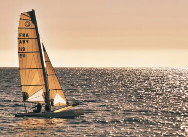 Catamarano a vela sul mare al tramonto, con vele spiegate e riflessi dorati sull'acqua, simbolo di avventura e libertà in mare.