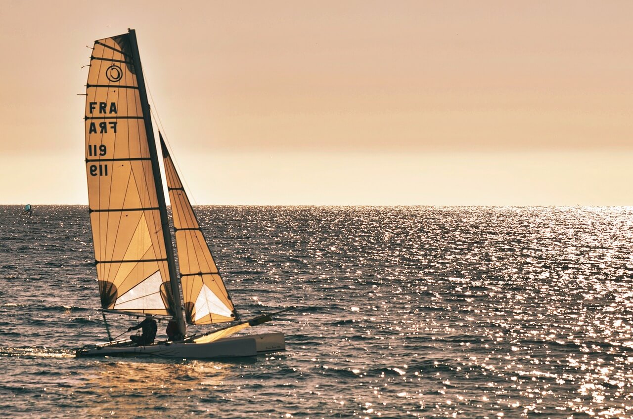 Catamarano a vela sul mare al tramonto, con vele spiegate e riflessi dorati sull'acqua, simbolo di avventura e libertà in mare.