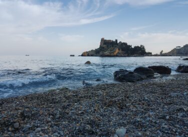 Spiaggia di ciottoli con onde leggere, rocce scure in primo piano e isolotto roccioso vegetato sullo sfondo, sotto un cielo variabile.