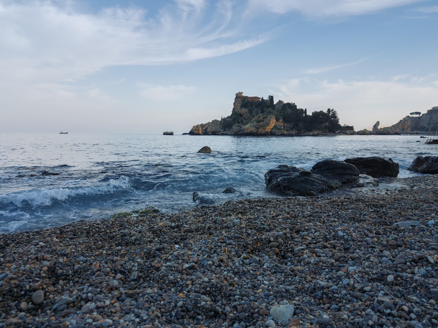 Spiaggia di ciottoli con onde leggere, rocce scure in primo piano e isolotto roccioso vegetato sullo sfondo, sotto un cielo variabile.