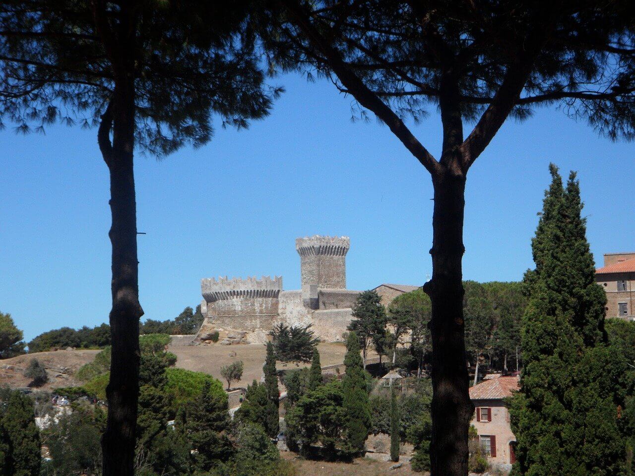 Vista del castello di Populonia medievale attraverso alberi in Toscana. Cielo sereno, architettura storica, natura rigogliosa.