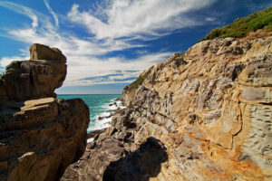 Scogliera rocciosa a Buca delle Fate, Piombino, con formazioni di arenaria erosa. Il mare turchese si infrange sulle rocce sotto un cielo blu con nuvole.