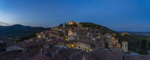 Borgo toscano al tramonto con tetti in cotto, colline verdi sullo sfondo e luci accese sulle case in primo piano