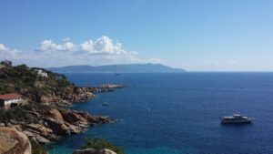 Costa rocciosa dell'isola del giglio con case affacciate sul mare azzurro e barca ancorata, isola visibile all’orizzonte sotto un cielo