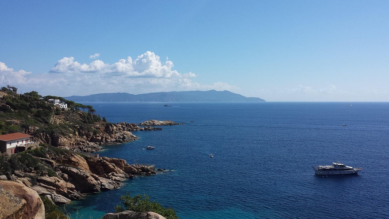 Costa rocciosa dell'isola del giglio con case affacciate sul mare azzurro e barca ancorata, isola visibile all’orizzonte sotto un cielo